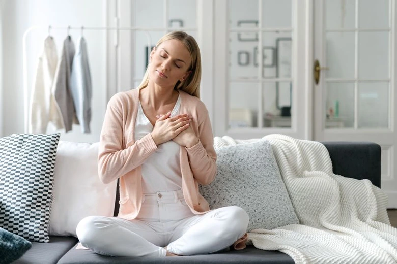 Žena sedí na gauči s zavřenýma očima při meditaci pro sebevědomí.