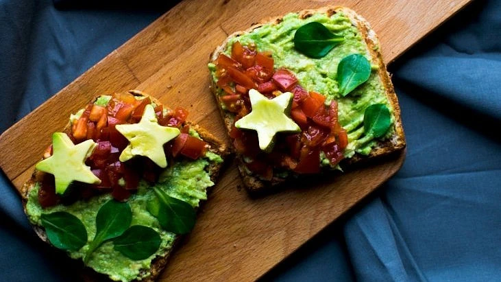 Dva kousky avokádového toastu s rajčaty a bazalkou na dřevěném talíři.