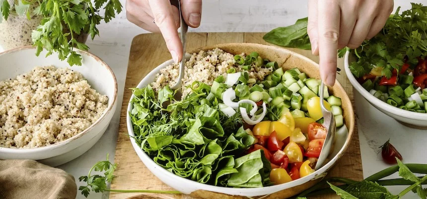 Rukou se míchá veganský salát s quinoou a zeleninou v dřevěné mísce.