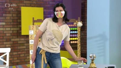 Žena v roušku stojí ve studiu před barevnou zdí.