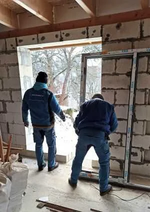 Dva dělníci instalují velké okno do zděné stěny.