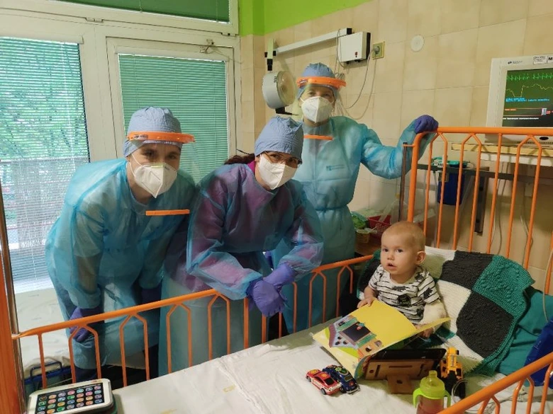 Tým lékařů v ochranném oblečení stojí u postele dítěte s hračkami.