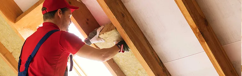 Muž ručně instaluje tepelnou izolaci mezi trámy v půdě.