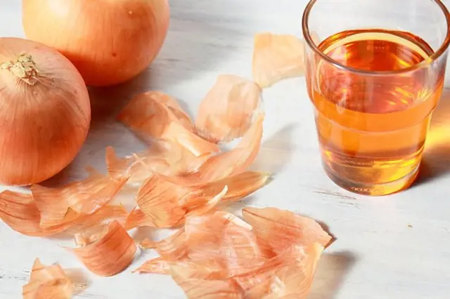 Cibule, cibulové slupky a voda s infuzí v sklenici na světlém dřevěném povrchu.