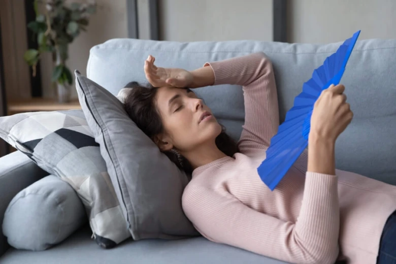 Žena leží na gauči s ventilátorem kvůli horku.