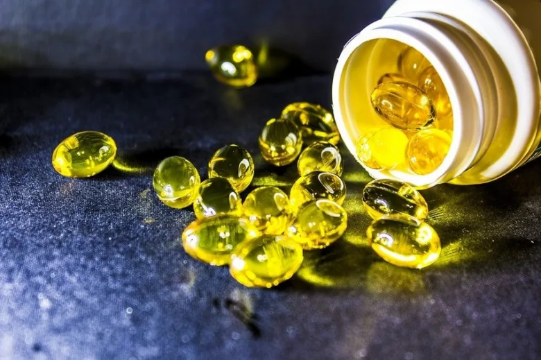 Žluté vitamínové tobolky vylije z bílé lahvičky na tmavý povrch.