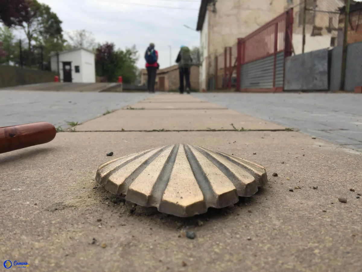 Kamenný mušlovitý symbol na cestě svatojakubské s poutníky v pozadí.