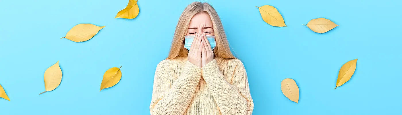 Žena v roušku kašle na modrém pozadí s podzimními listy.