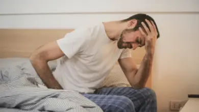 Muž sedí v posteli s bolestmi hlavy a zad.