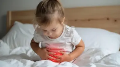 Malá dívka sedí na posteli a drží si břicho, což naznačuje střevní chřipku.